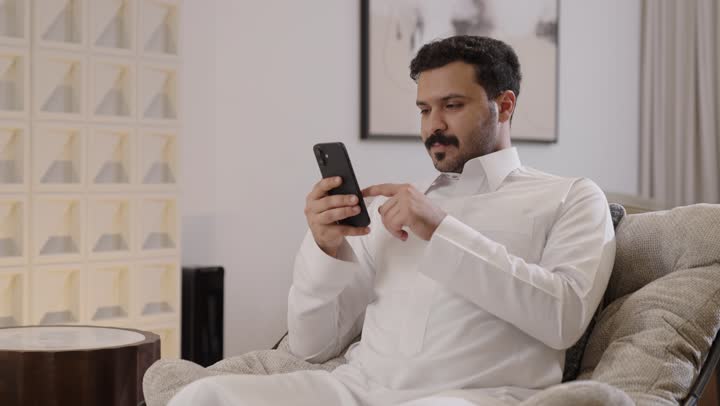 إرسال الرسائل النصية والصوتية عبر الجوال، رجل عربي خليجي سعودي مبتسم يرتدي الثوب التقليدي يمسك الهاتف المحمول بيده ينظر الى الكاميرا، تصفح وسائل التواصل الاجتماعي عبر الهاتف