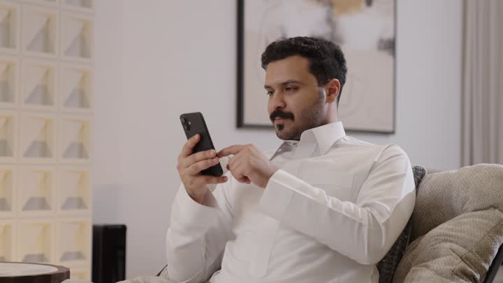 استخدام جهاز تقنى حديث، رجل عربي خليجي سعودي مبتسم يرتدي الثوب التقليدي يمسك الهاتف المحمول بيده ينظر الى الكاميرا، تصفح وسائل التواصل الاجتماعي عبر الجوال