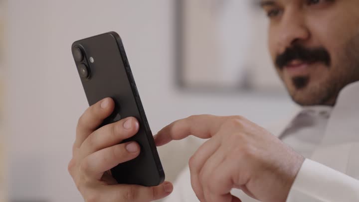 رجل عربي خليجي سعودي مبتسم يرتدي الثوب التقليدي يمسك الهاتف المحمول بيده، تصفح وسائل التواصل الاجتماعي عبر الجوال, استخدام جهاز تقنى حديث