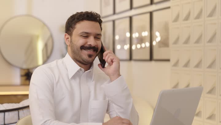 Chatting with family and friends, making a phone call using a mobile phone, an Arab Gulf Saudi man wearing traditional attire using a mobile phone in front of a laptop screen, having a good time.