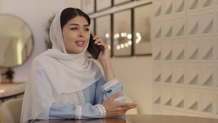 الاطمئنان على الاهل، امراة عربية خليجية سعودية محجبة ترتدى عباءة باللون الازرق الفاتح تقوم بالتحدث عبر الهاتف المحمول، التواصل مع الاصدقاء، استخدام جهاز تقنى حديث