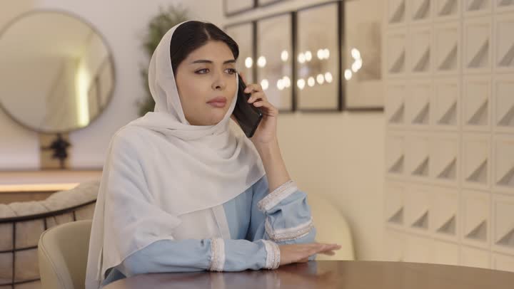 الدردشة عبر الجوال، امراة عربية خليجية سعودية محجبة ترتدى عباءة باللون الازرق الفاتح تقوم بالتحدث عبر الهاتف المحمول، التواصل مع الاصدقاء، استخدام جهاز تقنى حديث