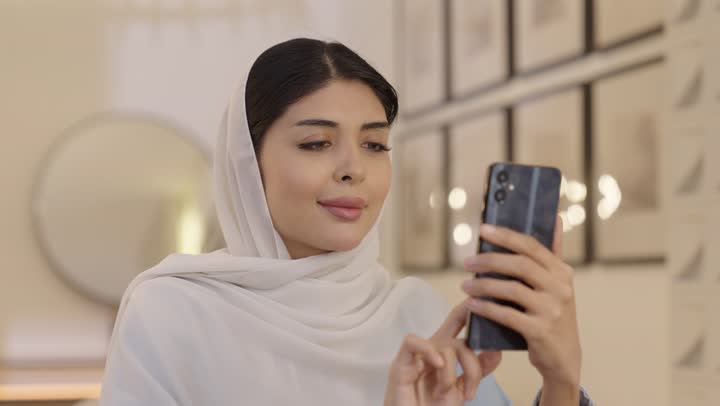 امراة عربية خليجية سعودية محجبة ترتدى عباءة باللون الازرق الفاتح تجلس فى غرفه المعيشة تقوم بتصفح الانترنت عبر الهاتف المحمول، قضاء وقت ممتع في المنزل، استخدام التقنية الحديثة