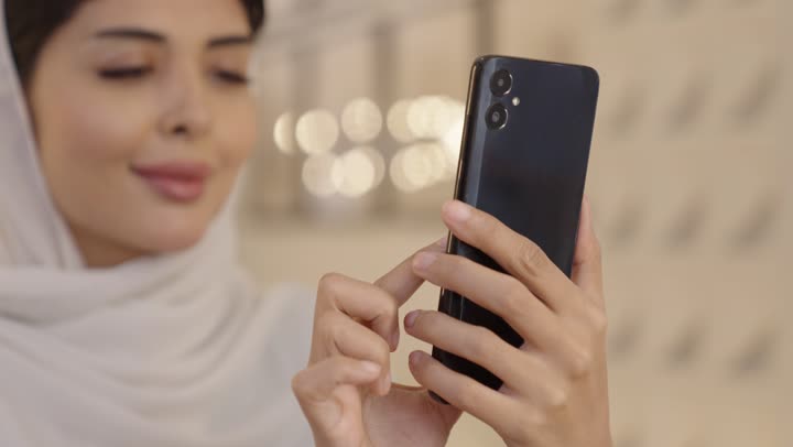 Checking on family and friends, an Arab Gulf Saudi woman wearing a light blue hijab and abaya is tapping on her mobile phone, shopping online.