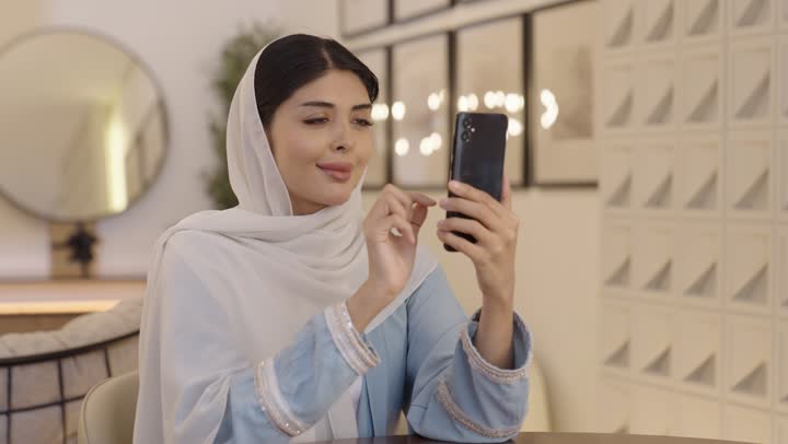 النظر الى الكاميرا بايماءات السرور، الاطمئنان على الاهل والاصدقاء، امرأة عربية خليجية سعودية ترتدى الحجاب وعباءة باللون الازرق الفاتح تقوم بالنقر على الهاتف المحمول، التسوق عبر الانترنت