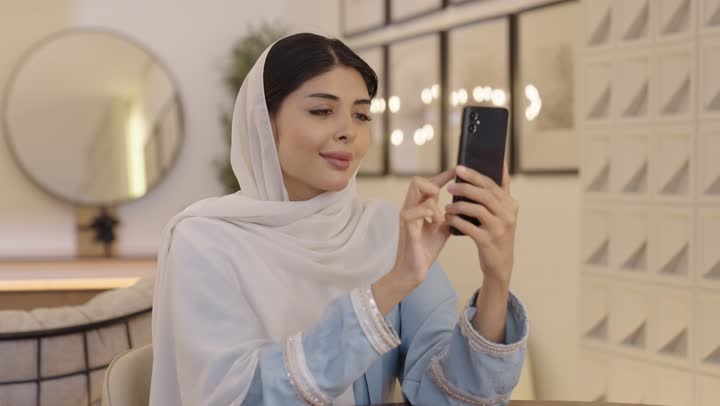 قضاء وقت ممتع في المنزل، الاطمئنان على الاهل والاصدقاء، امرأة عربية خليجية سعودية ترتدى الحجاب وعباءة باللون الازرق الفاتح تقوم بالنقر على الهاتف المحمول، التسوق عبر الانترنت