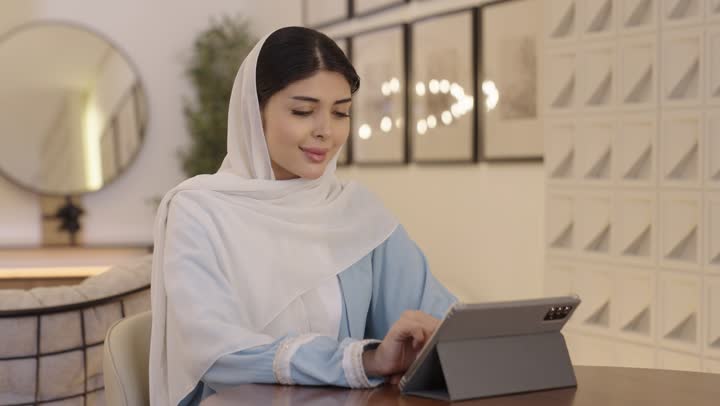 Online shopping, checking on family and friends, an Arab Gulf Saudi woman wearing a hijab and a light blue abaya browsing the internet on a tablet, chatting online.