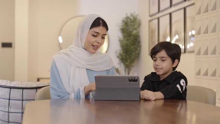 Integrating learning with technology, following online educational platforms, an Arab Gulf Saudi mother wearing a hijab and a light blue abaya sits with her son holding a tablet in her hand.
