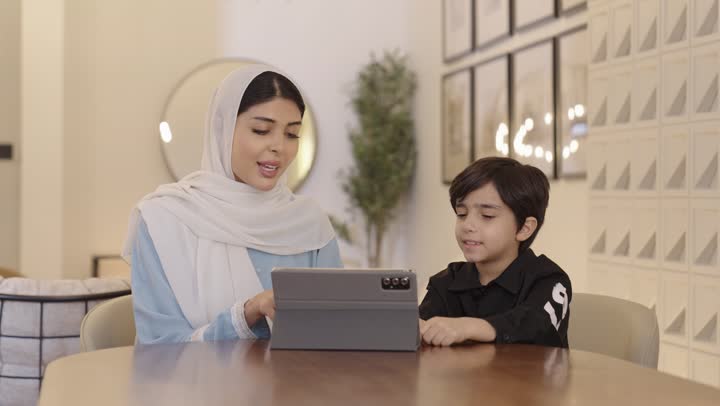 استخدام الاجهزه الحديثة و التقنية، قضاء وقت ممتع مع الابن، أم عربيه خليجيه سعودية ترتدي الحجاب وعباءة باللون الازرق الفاتح تجلس برفقة ولدها تمسك بيدها جهاز التابليت المحمول