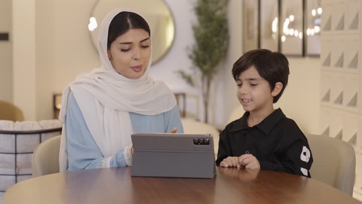 قضاء وقت ممتع مع الابن، استخدام الاجهزه الحديثة و التقنية، أم عربيه خليجيه سعودية ترتدي الحجاب وعباءة باللون الازرق الفاتح تجلس برفقة ولدها تمسك بيدها جهاز التابليت المحمول