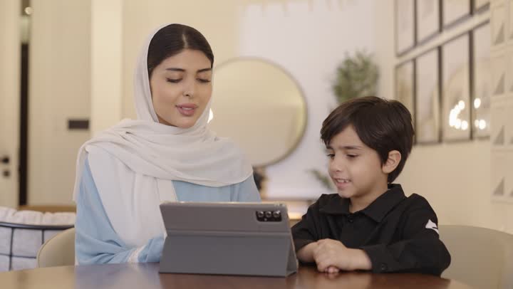 The trust in a mother's tenderness and her role in the lives of her children, the use of modern devices and technology, an Arab Gulf Saudi mother wearing a hijab and a light blue abaya sitting with her son holding a portable tablet in her hand.
