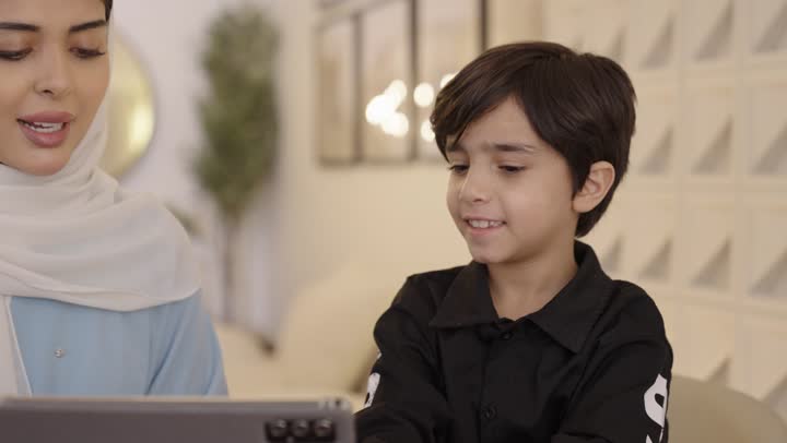 The concept of motherhood and childhood, the use of modern devices and technology, an Arab Gulf Saudi mother wearing a hijab and a light blue abaya sitting with her son holding a mobile tablet in her hand.