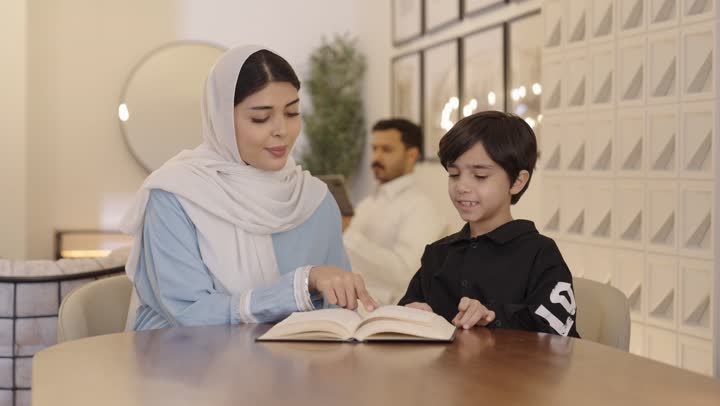 The passion for reading enjoyable stories and novels, the concept of education and knowledge, an Arab Gulf Saudi mother wearing a light blue hijab and abaya sitting with her son reading entertaining novels.