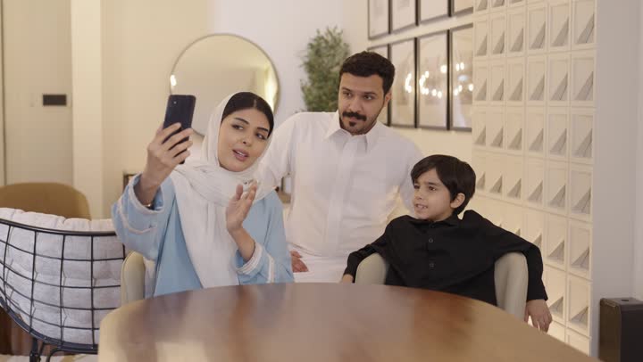 جو عائلي مرح، مفهوم الأسرة السعيدة، إستخدام أجهزة تقنية حديثة، عائلة خليجية سعودية تجلس على الأريكة تقوم باجراء مكالمة فيديو جماعيه عبر الانترنت باستخدام الهاتف