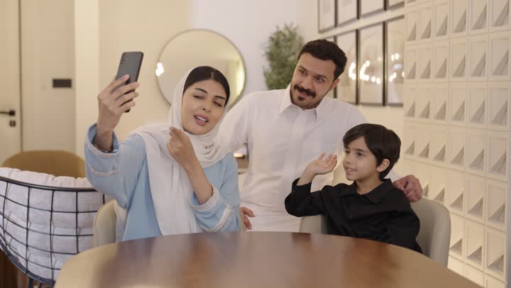 The concept of a happy family, cheerful family atmosphere, using modern technological devices, a Saudi Gulf family sitting on the couch making a group video call over the internet using a phone.