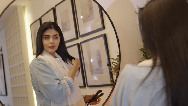 أمرأة عربيه خليجيه سعودية ترتدي عباءة بالون الازرق الفاتح تقوم بتصفيف شعرها امام المرأة باستخدام فرشاة الشعر، مفهوم العناية والاهتمام بالجمال والأناقة والمظهر الخارجي، تمشيط وترتيب الشعر، مفهوم الانوثه والنعومه