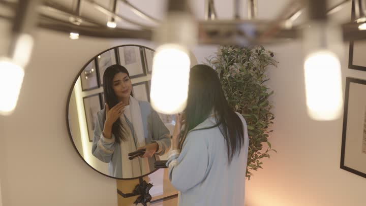 تمشيط وترتيب الشعر، مفهوم الانوثه والنعومه، مفهوم العناية والاهتمام بالجمال والأناقة والمظهر الخارجي، أمرأة عربيه خليجيه سعودية ترتدي عباءة بالون الازرق الفاتح تقوم بتصفيف شعرها امام المرأة باستخدام فرشاة الشعر