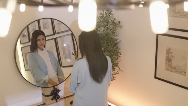 مفهوم الانوثه والنعومه ، تمشيط وترتيب الشعر، مفهوم العناية والاهتمام بالجمال والأناقة والمظهر الخارجي، أمرأة عربيه خليجيه سعودية ترتدي عباءة بالون الازرق الفاتح تقوم بتصفيف شعرها امام المرأة باستخدام فرشاة الشعر