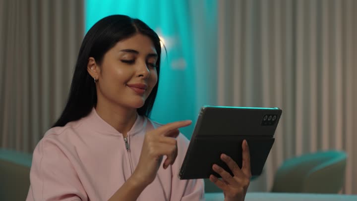 Browsing social media, a Gulf Arab Saudi woman wearing casual attire sits on the couch tapping on a mobile tablet, online shopping, completing tasks remotely, using modern and advanced devices and technologies.