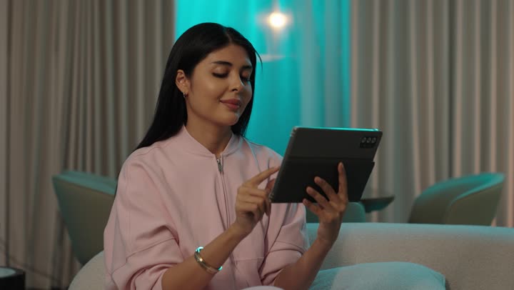 A Gulf Arab Saudi woman wearing casual attire is sitting on the sofa tapping on a mobile tablet, using tablet technology applications, online shopping, completing tasks remotely, and utilizing modern and advanced devices and technologies.