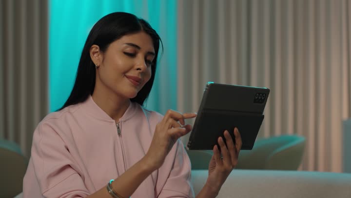 التطبيقات التقنية للأجهزة اللوحية، التسوق الالكتروني عبرالأونلاين، إنجاز المهام عن بعد، استخدام الأجهزة والتقنيات الحديثة والمتطورة، أمرأة عربيه خليجيه سعودية ترتدي الزى الكاجوال تجلس على الاريكة تقوم بالنقرعلى جهاز التابليت المحمول