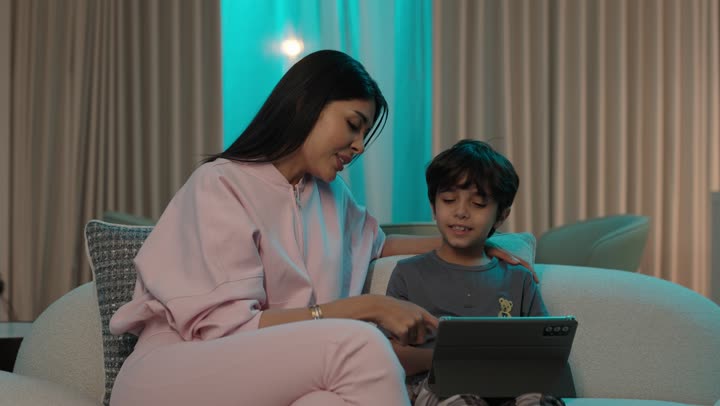 The trust in a mother's tenderness and her role in the lives of her children, the use of modern and advanced technological devices, an Arab Gulf Saudi mother dressed in casual attire sitting on the sofa tapping on a mobile tablet.