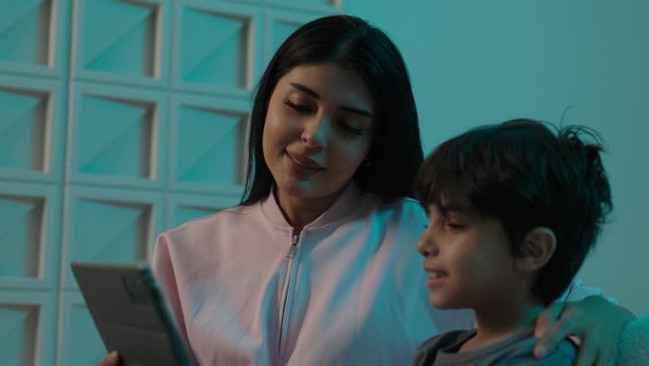 قضاء الوقت مع الابن، تشجيع الأولاد على التعلم والتثقف، تعزيز الروابط العائلية، مفهوم التربيه القائمه على الحب والحنان، أم عربيه خليجيه سعودية ترتدي الزى الكاجوال تجلس على الاريكة برفقة ولدها تقرأ الروايات عبر التابلت