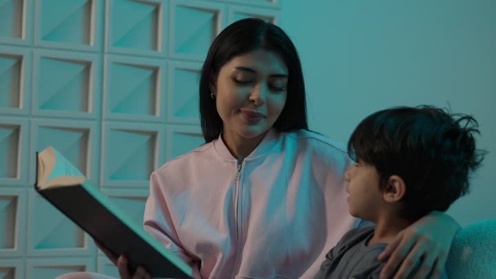 Encouraging children to learn and educate themselves, strengthening family bonds, spending time with the son, the concept of education based on love and affection, an Arab Gulf Saudi mother wearing casual attire sitting on the couch with her son reading entertaining novels.