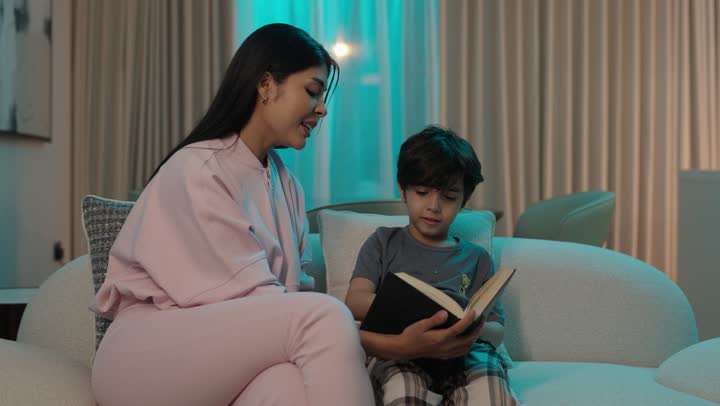 تعزيز الروابط العائلية، تشجيع الأولاد على التعلم والتثقف، قضاء الوقت مع الابن، مفهوم التربيه القائمه على الحب والحنان، أم عربيه خليجيه سعودية ترتدي الزى الكاجوال تجلس على الاريكة برفقة ولدها تقرأ الروايات المسليه