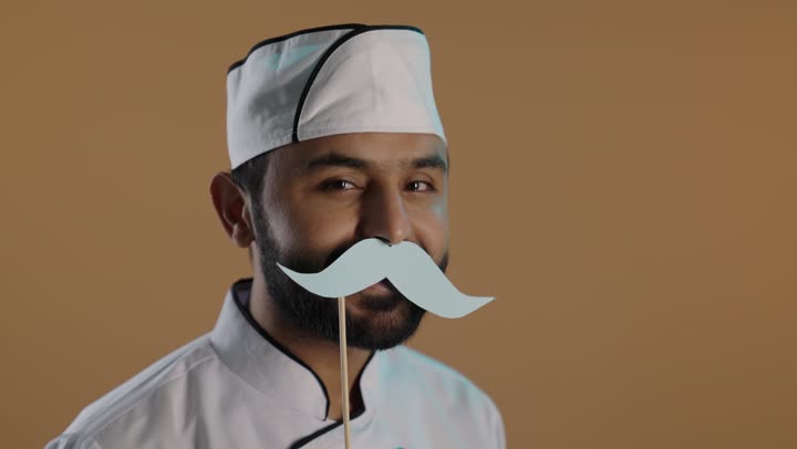 النظر الى الكاميرا بايماءات السعادة والسرور, الاحتفال السنوي باليوم العالمي للرجال ١٩ نوفمبر، بورتريه لرجل عربى خليجي سعودى يرتدي الزي الرسمي الخاص بمزاولة مهنة الطباخ يمسك بيده عود خشبي مع قصاصة ورقية على شكل شارب، خلفية بنية