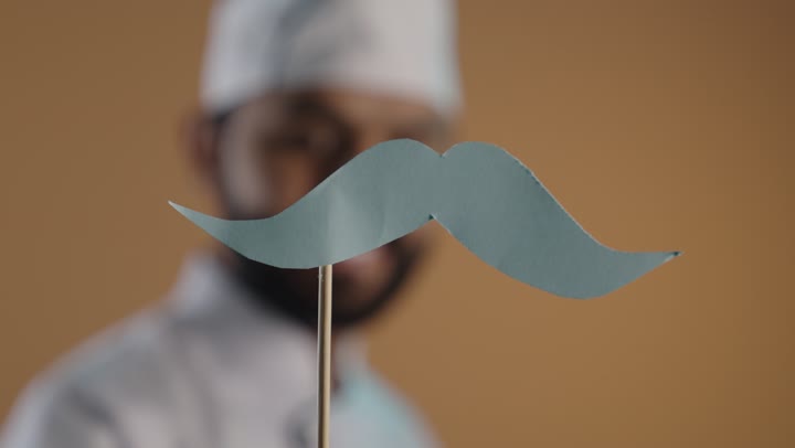 اليوم العالمي للرجل 19 نوفمبر، بورتريه  مقرب ليد رجل عربى خليجي سعودى يرتدي الزي الرسمي الخاص بمزاولة مهنة الطباخ يمسك بيده عود خشبي مع قصاصة ورقية على شكل شارب، خلفية بنية