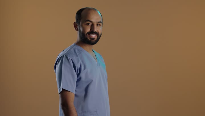 الوقوف بأيدى متقاطعة، مهن ووظائف طبية، الاهتمام بصحة المرضى والقيام بالرعاية الصحية, بورتريه من الجانب لممرض عربي خليجي سعودي يرتدي اليونيفورم والقفازات ينظرالى الكاميرا ، خلفية بنية