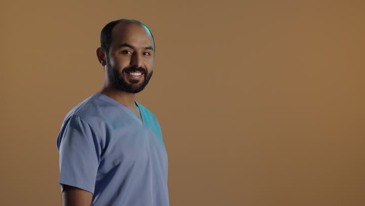 مهن ووظائف طبية، الاهتمام بصحة المرضى والقيام بالرعاية الصحية, بورتريه من الجانب لممرض عربي خليجي سعودي يرتدي اليونيفورم والقفازات ينظر الى الكاميرا بايدى متقاطعة، خلفية بنية