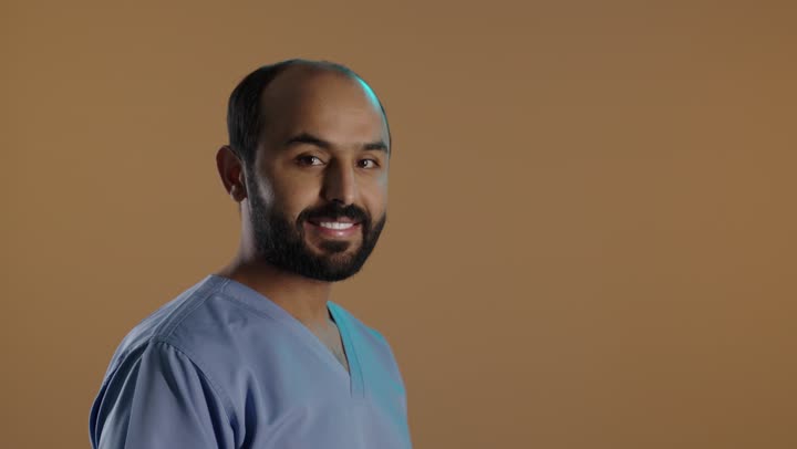 الاهتمام بصحة المرضى والقيام بالرعاية الصحية, بورتريه من الجانب لممرض عربي خليجي سعودي يرتدي اليونيفورم والقفازات ينظر الى الكاميرا بايدى متقاطعة، مهن ووظائف طبية، خلفية بنية