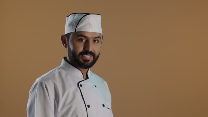 Standing upright with crossed arms, a portrait of a smiling Saudi Arabian Gulf man wearing the official uniform of a chef, looking at the camera with expressions of happiness and joy, with a brown background.