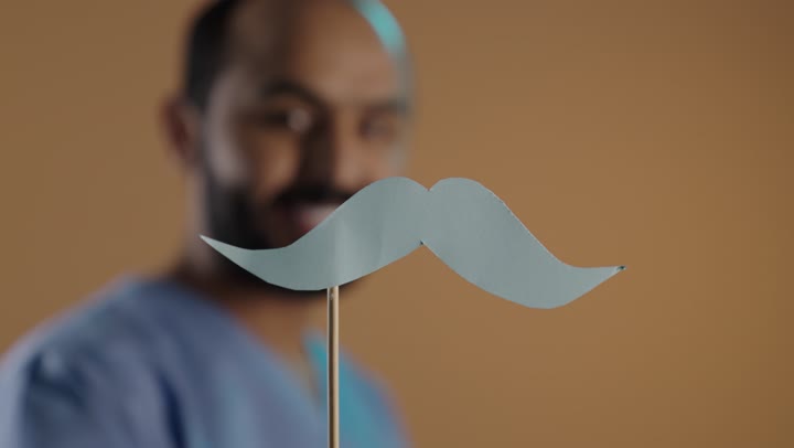 اليوم العالمي للرجل 19 نوفمبر، ايماءات السعادة والسرور، العمل في القطاع الصحي ومفهوم مهنة الطب، بورتريه من الجانب لممرض عربي خليجي سعودي يرتدي اليونيفورم والقفازات يمسك بيده عود خشبي مع قصاصة ورقية على شكل شارب، خلفية بنية