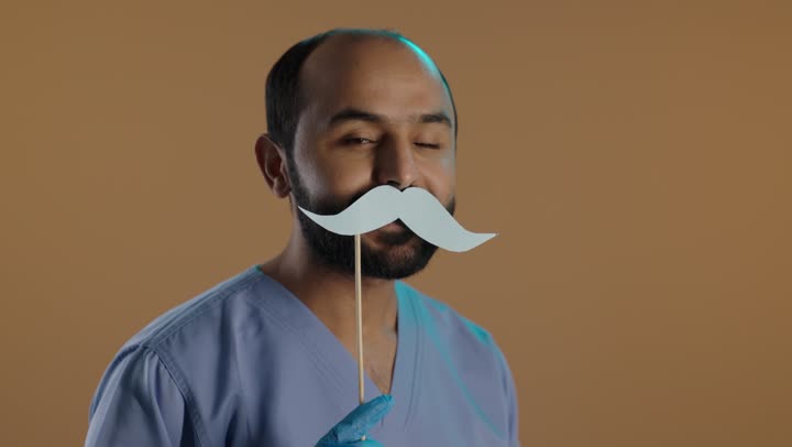 Working in the health sector and the concept of the medical profession, International Men's Day, November 19, a side portrait of a Saudi Gulf Arab nurse wearing a uniform and gloves holding a wooden stick with a paper cutout in the shape of a mustache, gestures of happiness and joy, brown background.