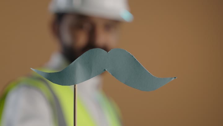 الاحتفال بالمناسبات العالمية، بورتريه لمهندس عربي خليجي سعودى يرتدي الثوب التقليدي والستره الوقائيه ويضع خوذه الحماية على رأسه يمسك بيده عود خشبي مع قصاصة ورقية على شكل شارب، اليوم العالمي للرجل 19 نوفمبر، خلفية بنية