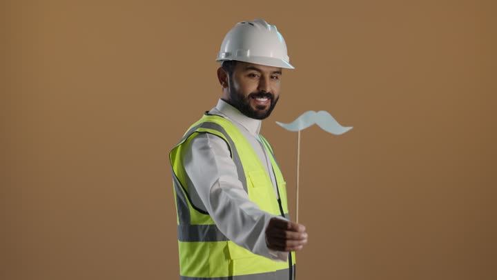 بورتريه لمهندس عربي خليجي سعودى يرتدي الثوب التقليدي والستره الوقائيه ويضع خوذه الحماية على رأسه يمسك بيده عود خشبي مع قصاصة ورقية على شكل شارب، الاحتفال بالمناسبات العالمية، اليوم العالمي للرجل 19 نوفمبر، خلفية بنية