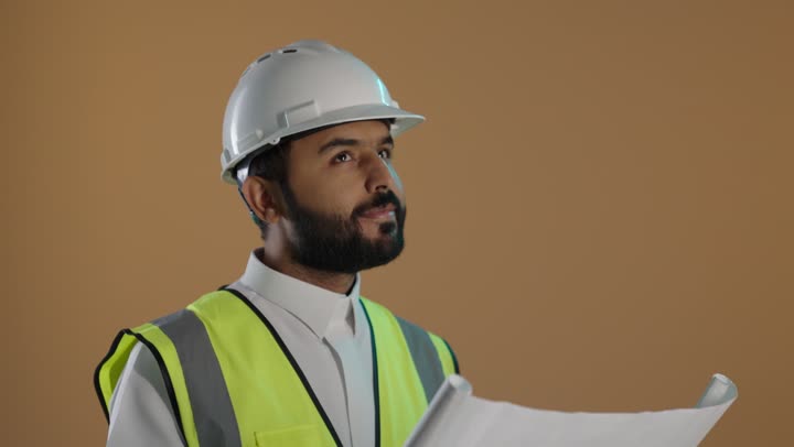 A portrait of a Saudi Arabian Gulf engineer wearing traditional attire and a safety vest, with a protective helmet on his head, studying and browsing engineering plans, examining and scrutinizing construction plans, the concept of engineering and construction work, development and follow-up of engineering projects, with a brown background.