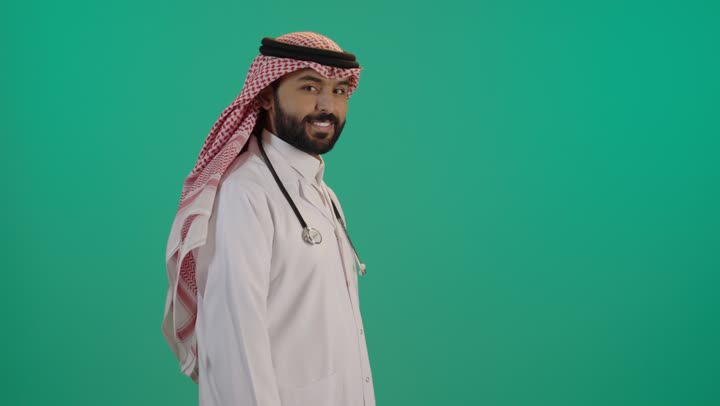 بورتريه مقرب لطبيب عربي خليجي سعودي مبتسم يرتدي الثوب التقليدي والشماغ مع المعطف الأبيض يضع سماعة الفحص ينظر إلى الكاميرا بتعابير السعادة والسرور، الوقوف باستقامة بايماءات ايدي متقاطعة, خلفية خضراء