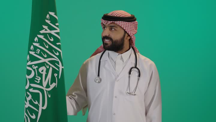 The anniversary of the unification of the Kingdom of Saudi Arabia, a portrait of an Arab Gulf Saudi doctor wearing traditional attire and a white coat with a stethoscope holding the Kingdom's flag, commemorating Saudi National Day on September 23, with a green background.