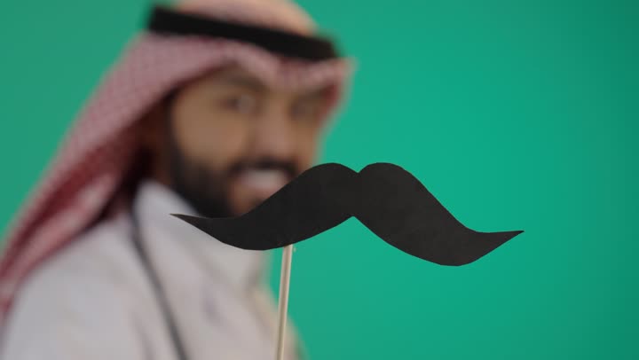 الاحتفال بالمناسبات العالمية، اليوم العالمي للرجل 19 نوفمبر، بورتريه مقرب ليد رجل عربي خليجي سعودي يرتدي الثوب التقليدي والشماغ ويمسك عود خشبي مع قصاصة ورقية على شكل شارب، الاهتمام بصحة الرجل، خلفية خضراء