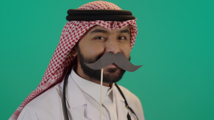 الإهتمام بمظهر الخارجي، اليوم العالمي للرجل 19 نوفمبر، الإحتفال بالمناسبات العالمية، الحفاظ على صحة الذكور، بورتريه لرجل عربي خليجي سعودي يرتدي الثوب التقليدي و الشماغ يمسك قصاصة ورقية على شكل شارب و يقربها من وجهه، خلفية خضراء