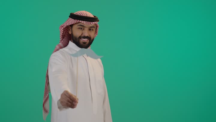 تعابير الرضا و الثقة بالنفس, اليوم العالمي للرجل 19 نوفمبر، بورتريه لرجل عربي خليجي سعودي يرتدي الثوب التقليدي والشماغ يمسك بيده عود خشبي مع قصاصة ورقية على شكل شارب، الإهتمام بصحة الرجال، ايماءات السعادة والسرور, خلفية خضراء