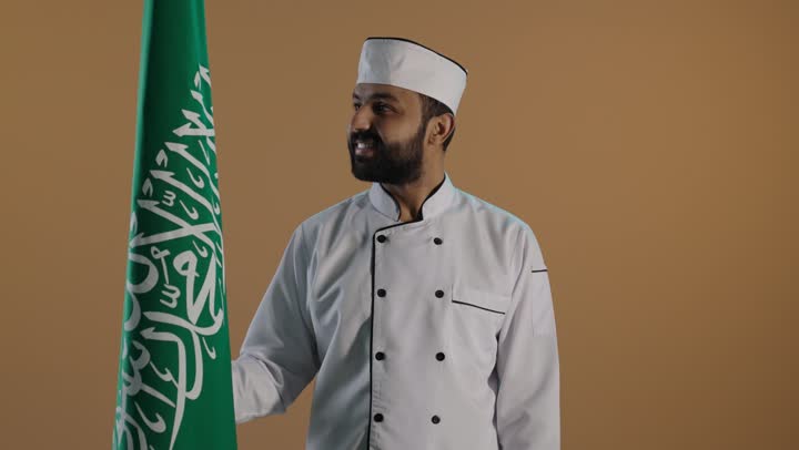 Expressing love for the homeland and belonging to it, a portrait of a Saudi Arabian Gulf chef wearing the official uniform for practicing the profession of cooking while holding the flag of the Kingdom, achieving growth and development through the efforts of the nation's sons, celebrating the anniversary of Saudi National Day on September 23 and the national holiday, with a brown background.