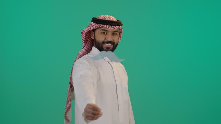 بورتريه مقرب لرجل عربي خليجي سعودي يرتدي الثوب التقليدي والشماغ يمسك بيده عود خشبي مع قصاصة ورقية على شكل شارب، تعابير الرضا و الثقة بالنفس, اليوم العالمي للرجل 19 نوفمبر، الإهتمام بصحة الرجال، ايماءات السعادة والسرور, خلفية خضراء