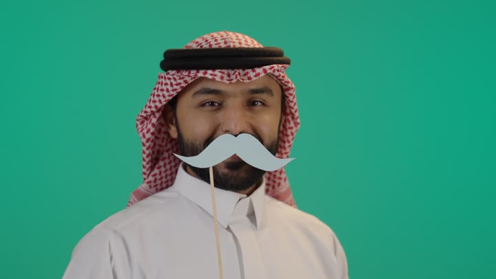 اليوم العالمي للرجل 19 نوفمبر، بورتريه مقرب لرجل عربي خليجي سعودي يرتدي الثوب التقليدي والشماغ يمسك بيده عود خشبي مع قصاصة ورقية على شكل شارب، تعابير الرضا و الثقة بالنفس, الإهتمام بصحة الرجال، ايماءات السعادة والسرور, خلفية خضراء