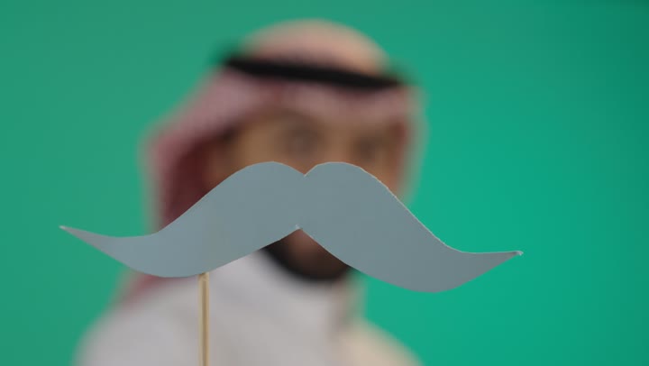 الاهتمام بالصحة الجسدية للذكور، اليوم العالمي للرجل 19 نوفمبر، بورتريه مقرب لرجل عربي خليجي سعودي يرتدي الثوب التقليدي والشماغ يمسك بيده عود خشبي مع قصاصة ورقية على شكل شارب, ايماءات السعادة والسرور, خلفية خضراء