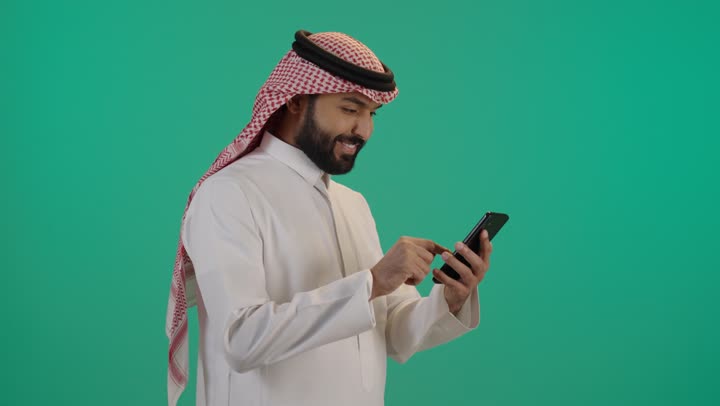 استخدام الهاتف المحمول لإنجاز المهام، تصفح وسائل التواصل الاجتماعي, استخدام الأجهزة التقنية الحديثة و المتطورة, رجل عربي خليجي سعودي مبتسم يرتدي الثوب التقليدي والشماغ يستخدم الجوال، خلفية خضراء